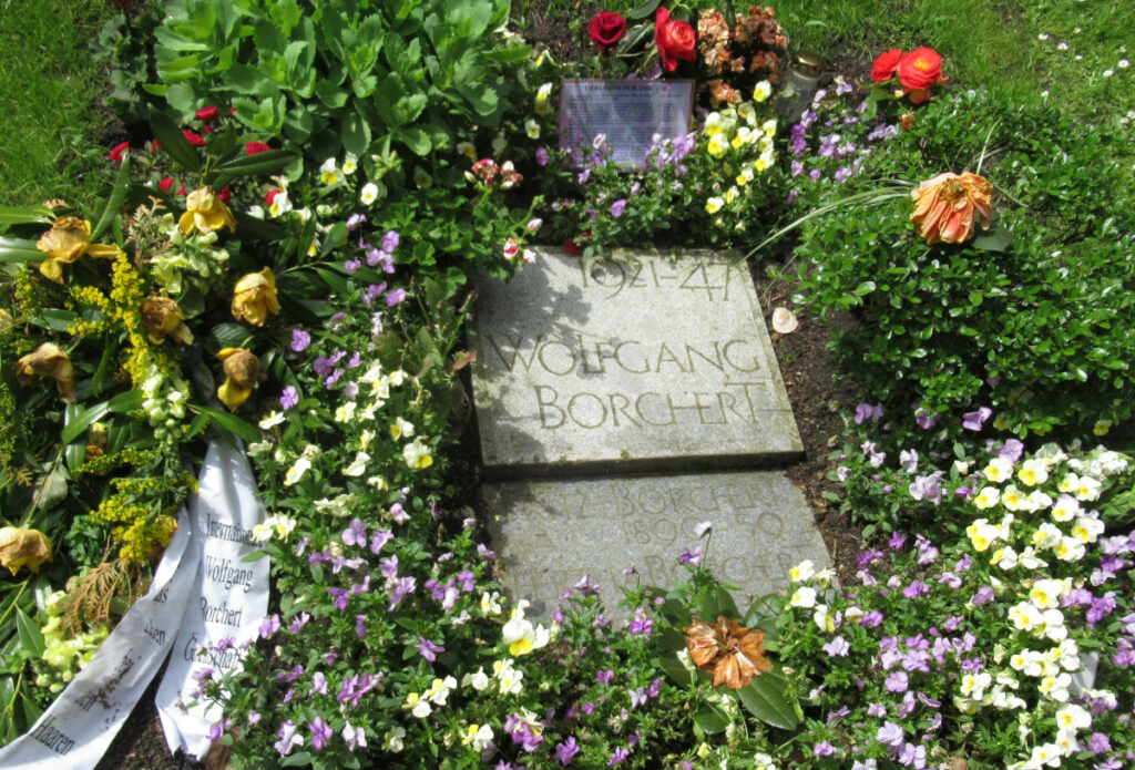 Zu sehen ist das Grab von Wolfgang Borchert, der im Mai 2021 seinen 100.Geburtstag feierte. Das Grab ist mit frischen Blumen geschmückt. Ein Kranz ist mit dem Zitat „Pack das Leben bei den Haaren“ beschriftet.