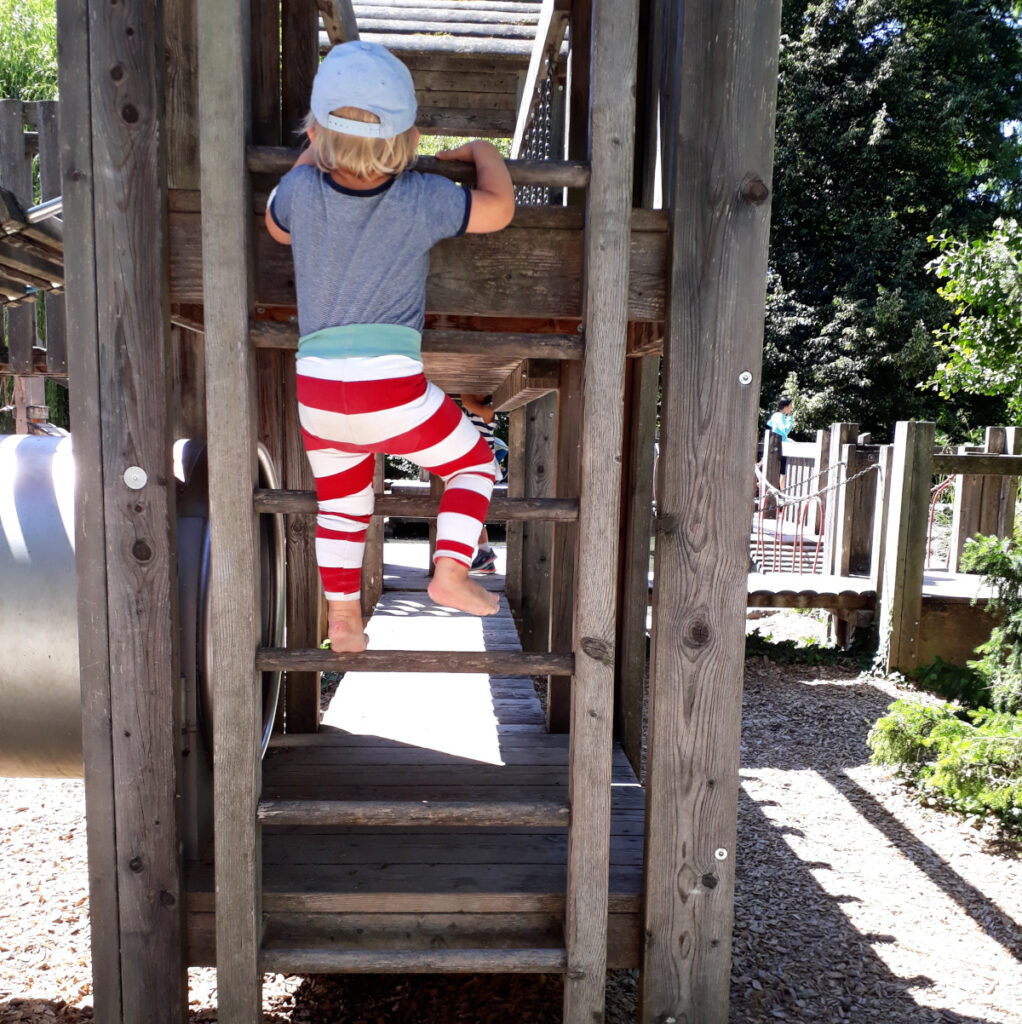 Es ist ein Kleinkind zu sehen, das an einem Holzspielgerät eine Leiter hinaufklettert.
