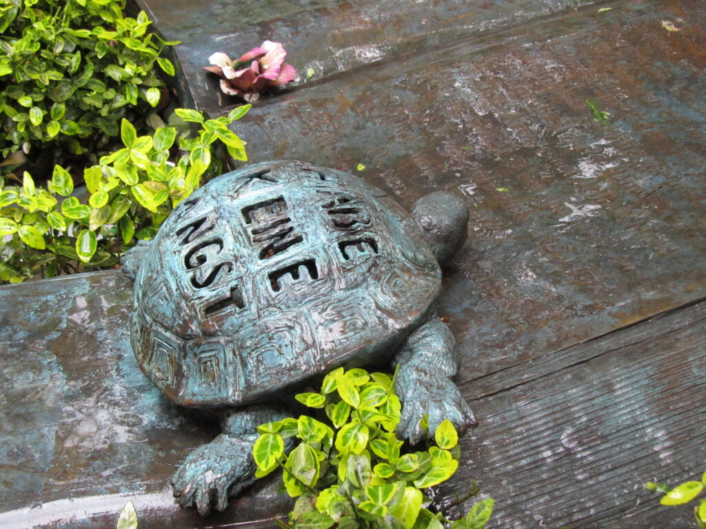 Waldfriedhof München, Detailaufnahme vom Grab des Schriftstellers Michael Ende. Eine Bronzeschildkröte, die die Worte „Habe Keine Angst“ auf ihrem Panzer trägt, in Anlehnung an die Romanfigur Kassiopeia aus seinem Roman „Momo“. Im Hintergrund sieht man ein aufgeschlagenes Buch aus Bronze und Grünpflanzen