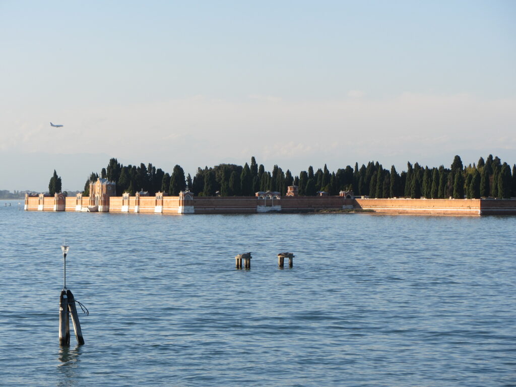 Friedhofsinsel San Michele, Venedig. Im Wasser liegt eine Insel, umschlossen von einer rötlichen Mauer. Einige ornamental verzierte Bögen und Türmchen sind zu erkennen. Dahinter Zypressen.
