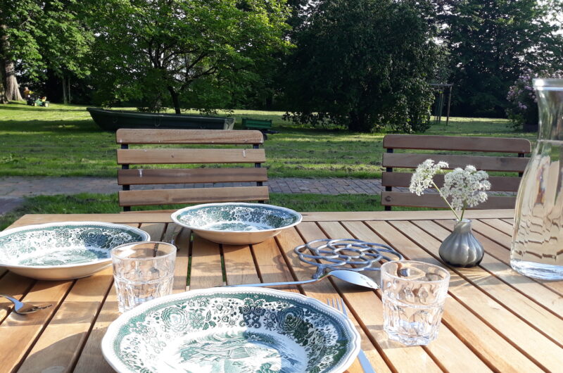 Zu sehen ist im Vordergrund ein gedeckter Tisch mit altem Villeroy und Boch-Geschirr, Giersch in einem Väschen und einer Wasserkaraffe. Im Hintergrund ein Boot und ein parkähnlicher Garten.