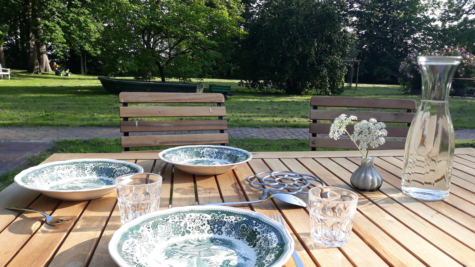 Zu sehen ist im Vordergrund ein gedeckter Tisch mit altem Villeroy und Boch-Geschirr, Giersch in einem Väschen und einer Wasserkaraffe. Im Hintergrund ein Boot und ein parkähnlicher Garten.