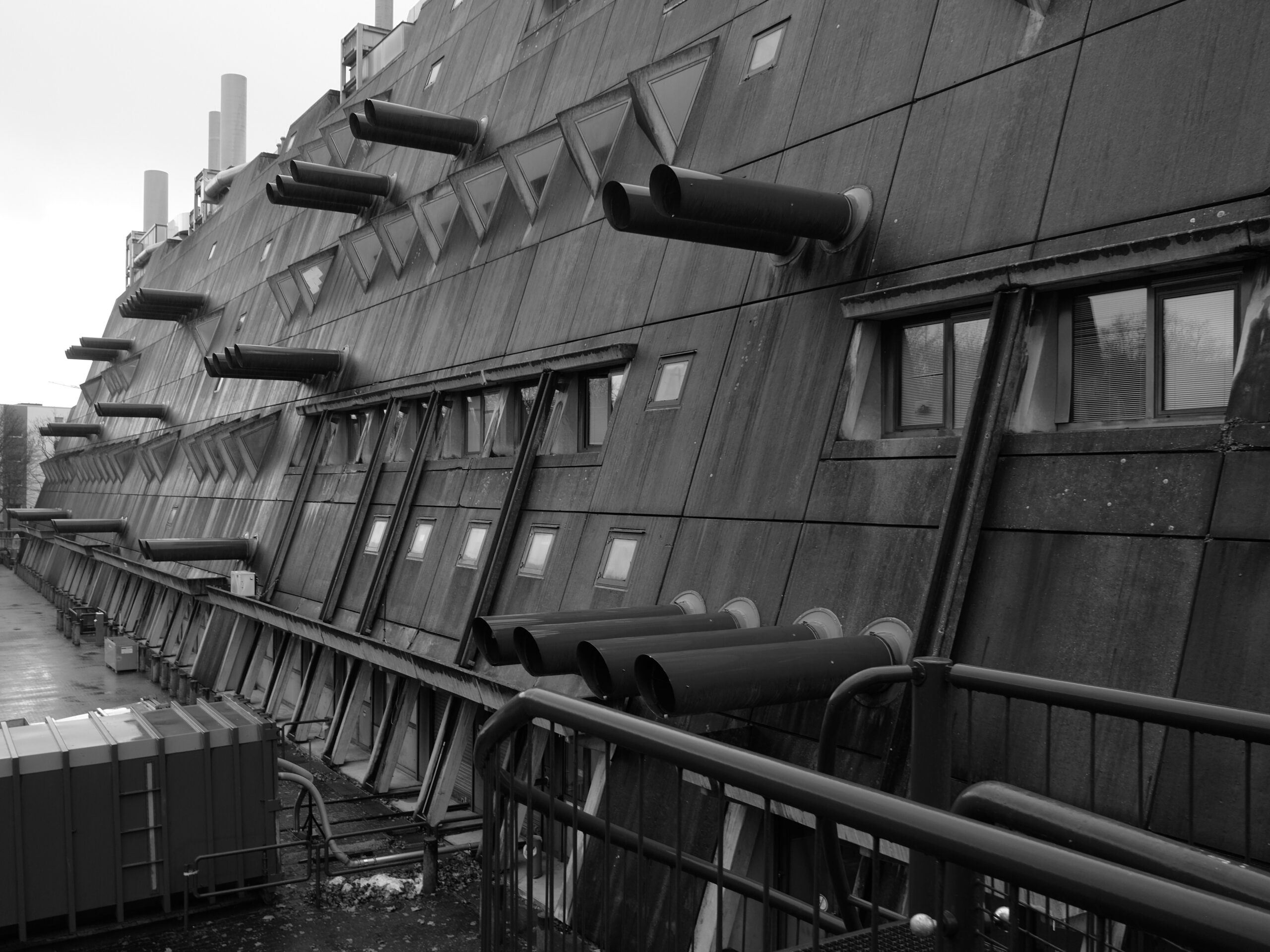 Eine Seite eines brutalistischen Gebäudes in Schwarz-Weiß. Lange Lüftungsrohre ragen aus der angeschrägten Beton-Fassade.