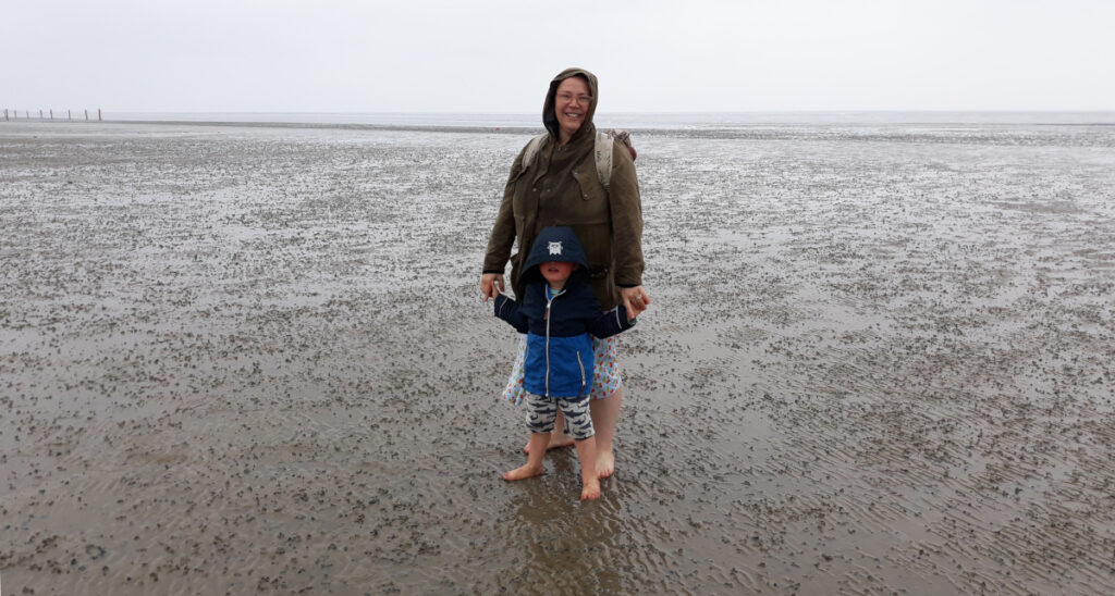 Eine Frau und ein Kleinkind stehen bei schlechtem Wetter im Watt. Der Hintergrund ist grau und diesig.