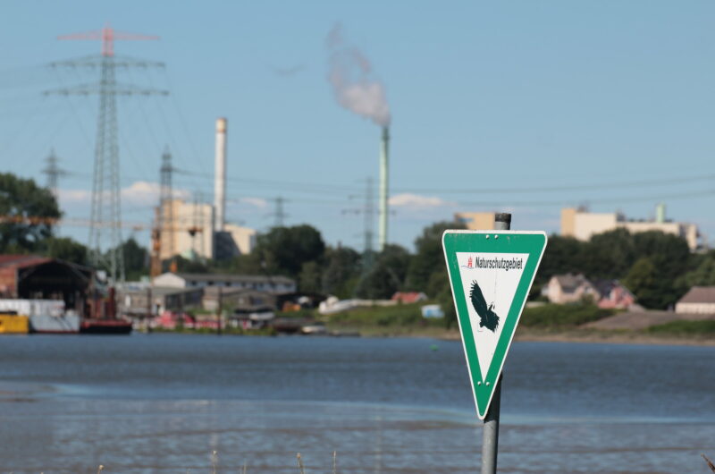 Ein Schild "Naturschutzgebiet" vor einer Wasserfläche. Im Hintergrund verschwommen eine Industrielandschaft