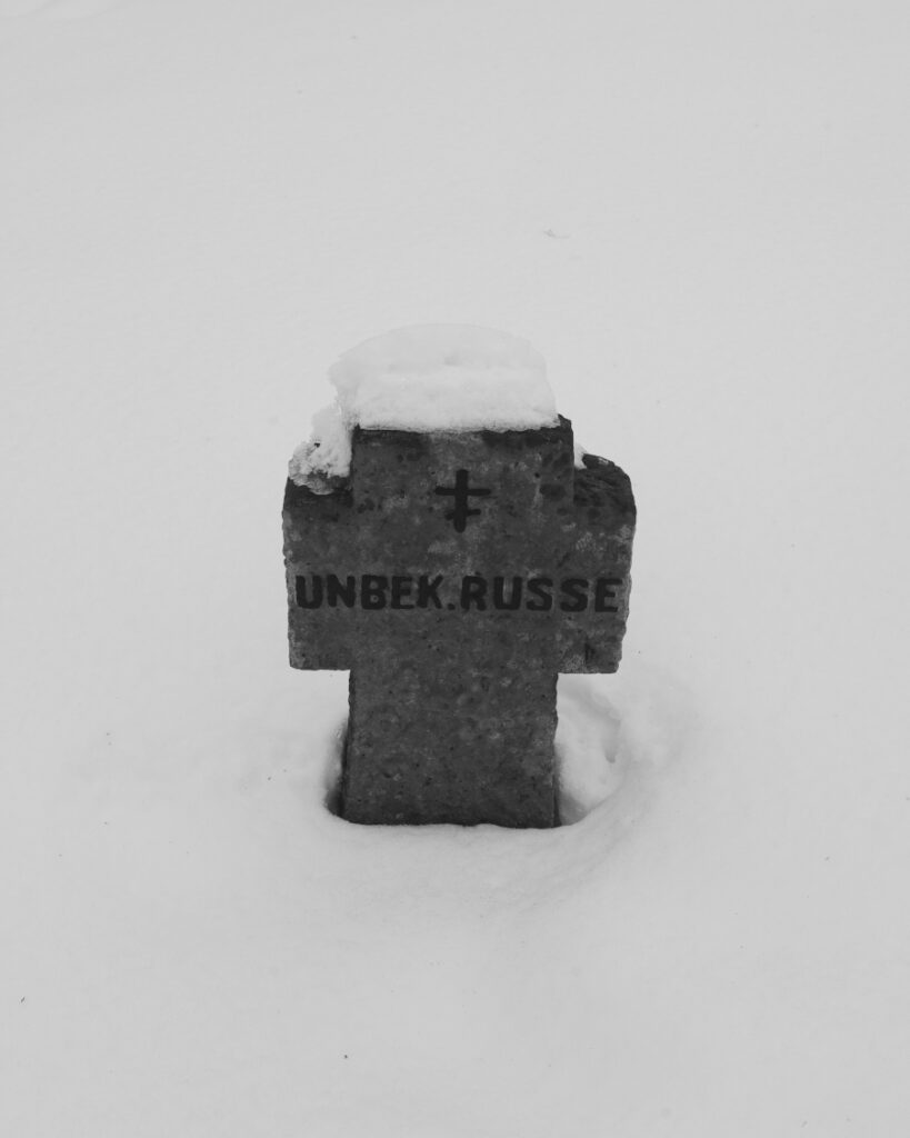 Schwarz-Weiss-Aufnahme von einem Grabstein, in Kreuzform, im Schnee mit der Inschrift „Unbekanter Russe“.