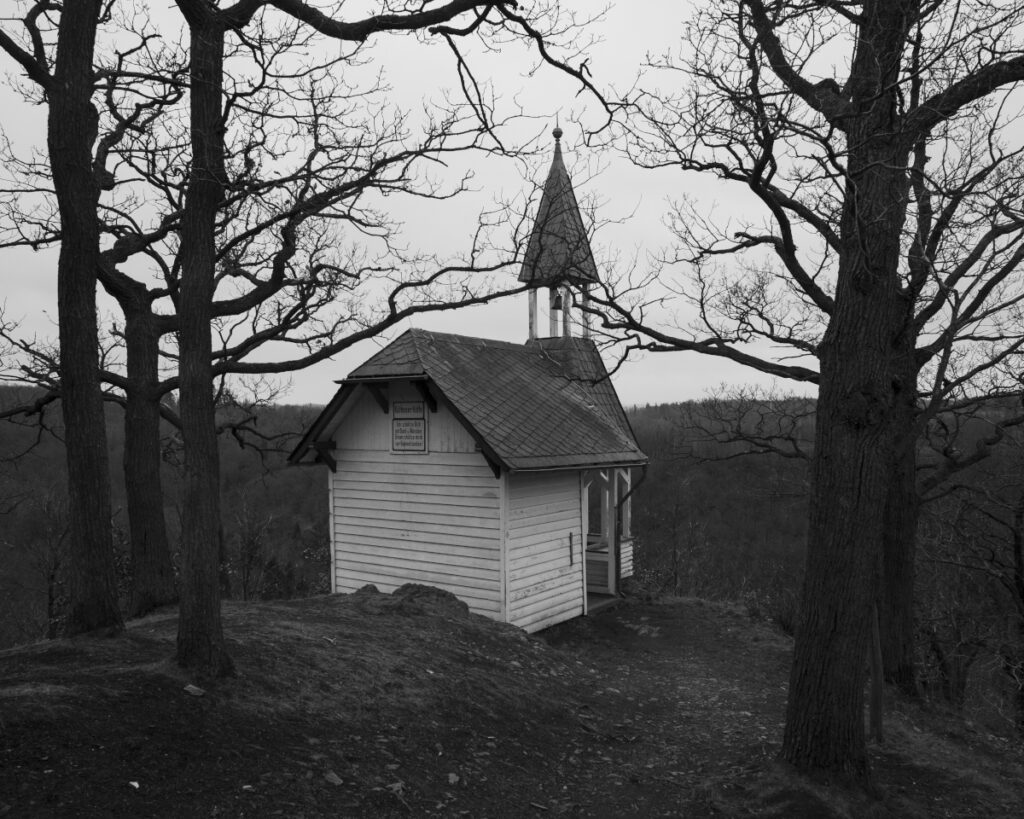 Schwarz-Weiss-Aufnahme von einer Kapelle ,die zwischen Bäumen auf dem Gipfel eines Berges steht.
