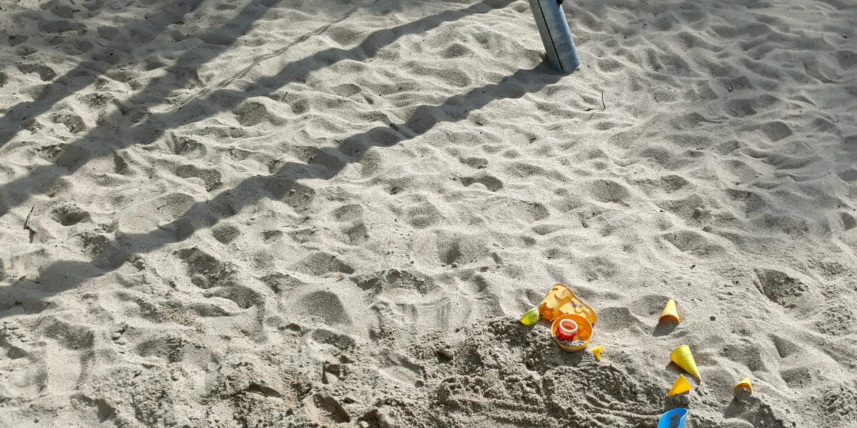 Ein Spielplatz mit viel Sand, im Vordergrund Sandspielzeug und der Sahtten einer Schaukel im Hintergrund