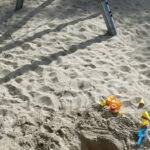 Ein Spielplatz mit viel Sand, im Vordergrund Sandspielzeug und der Sahtten einer Schaukel im Hintergrund