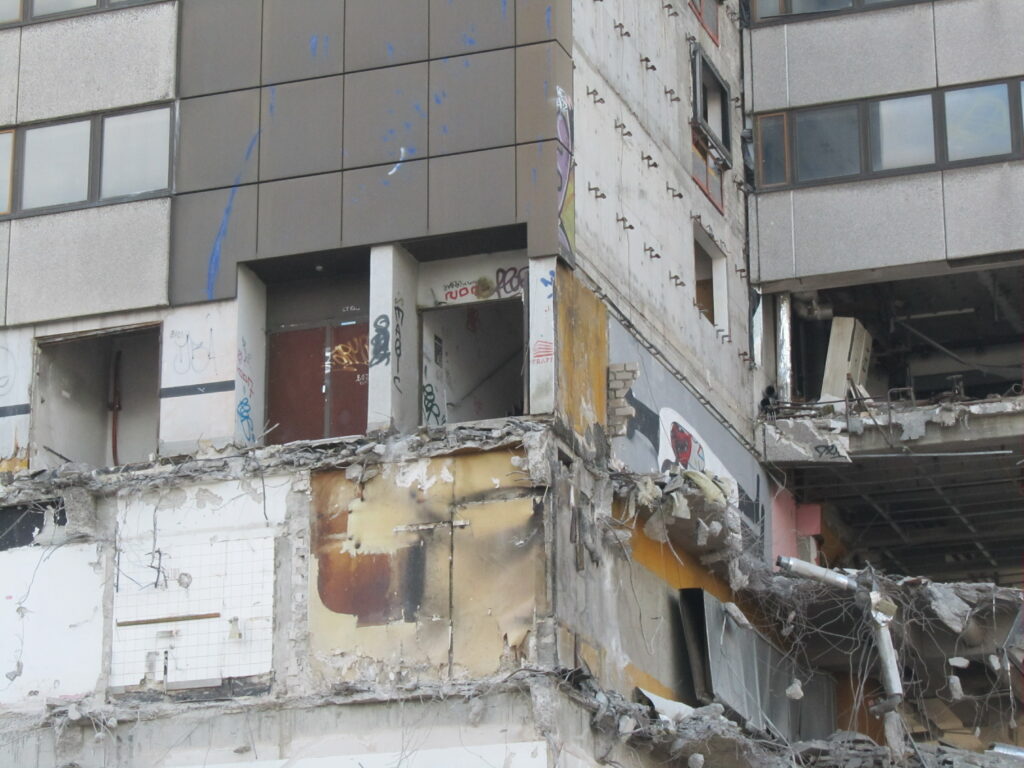 Man sieht ein Gebäude, das gerade abgerissen wird. Verschiedene Etagen sind zu sehen, Kabel hängen von den Wänden.