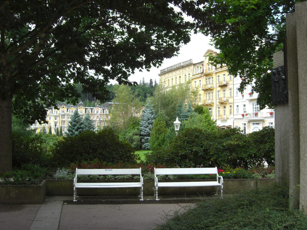 Zwei weiße Bänke umgeben von grünen Bäumen und Sträuchern. Im Hintergrund mehrgeschossige Altbauten.