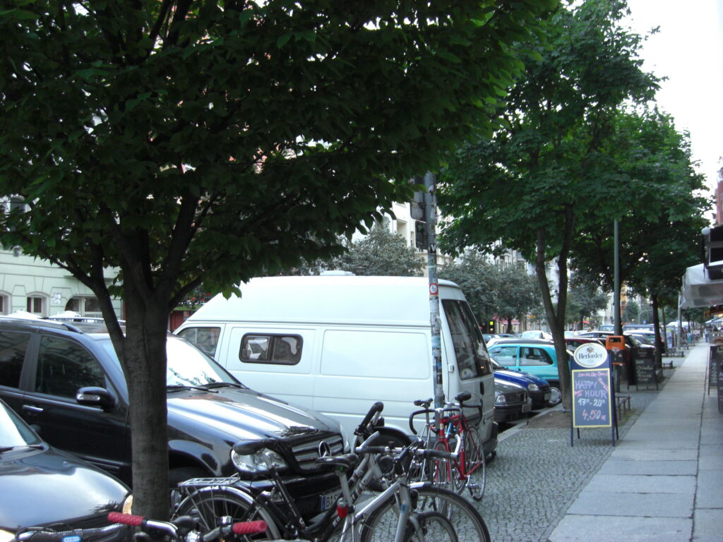 Ein schmaler Bürgersteig mit ein paar Fahrrädern, daneben sehr viele geparkte Autos.