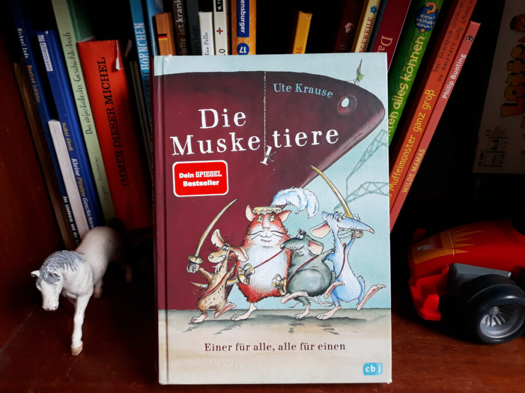 Das Buch „Die Muskeltiere“ von Ute Krause vor einem Regal und Spielzeugen