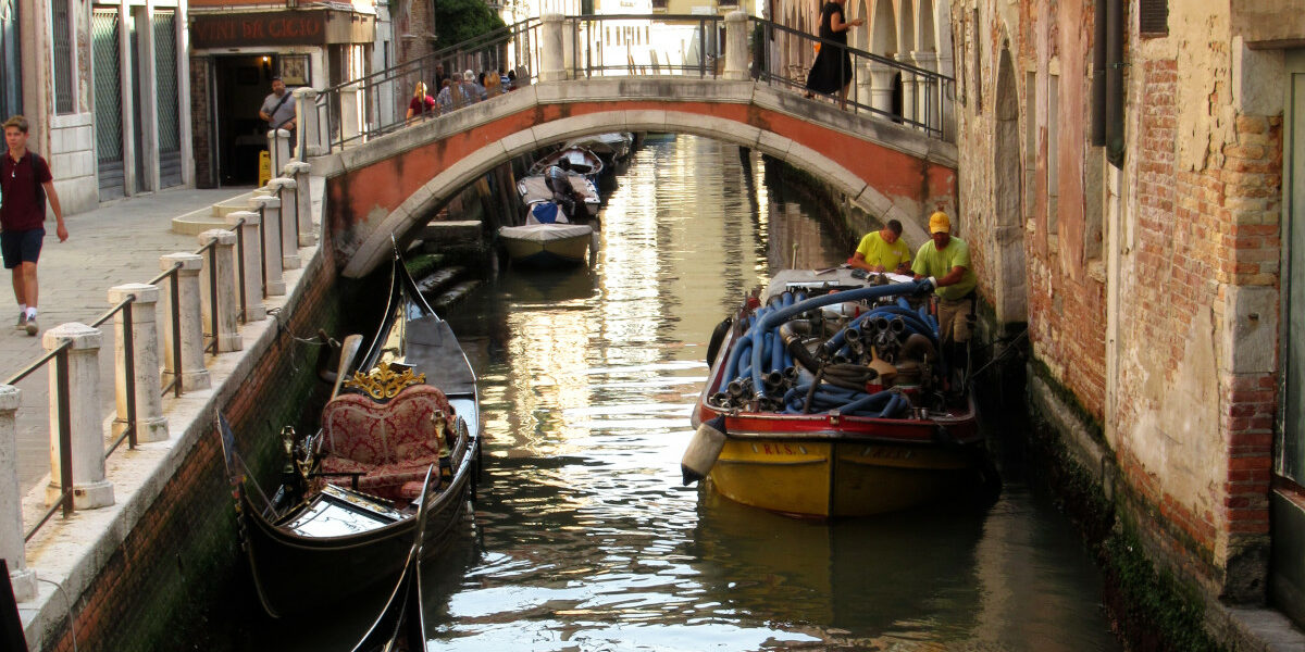 Ein Kanal in Venedig, linker Hand zwei historische Gondeln, rechts ein modernes Boot mit Baumaterial, im Hintergrund eine Brücke.