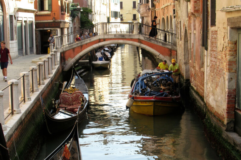 Ein Kanal in Venedig, linker Hand zwei historische Gondeln, rechts ein modernes Boot mit Baumaterial, im Hintergrund eine Brücke.