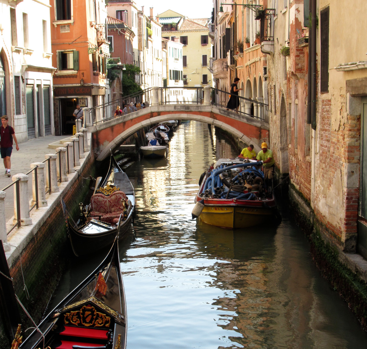 Ein Kanal in Venedig, linker Hand zwei historische Gondeln, rechts ein modernes Boot mit Baumaterial, im Hintergrund eine Brücke.