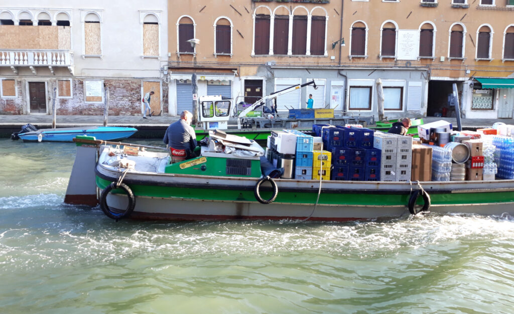Ein voll beladenes Transportboot fährt durch die Kanäle Venedigs. Im Hintergrund lädt ein Müllboot die Container der Müllarbeiter:innen.