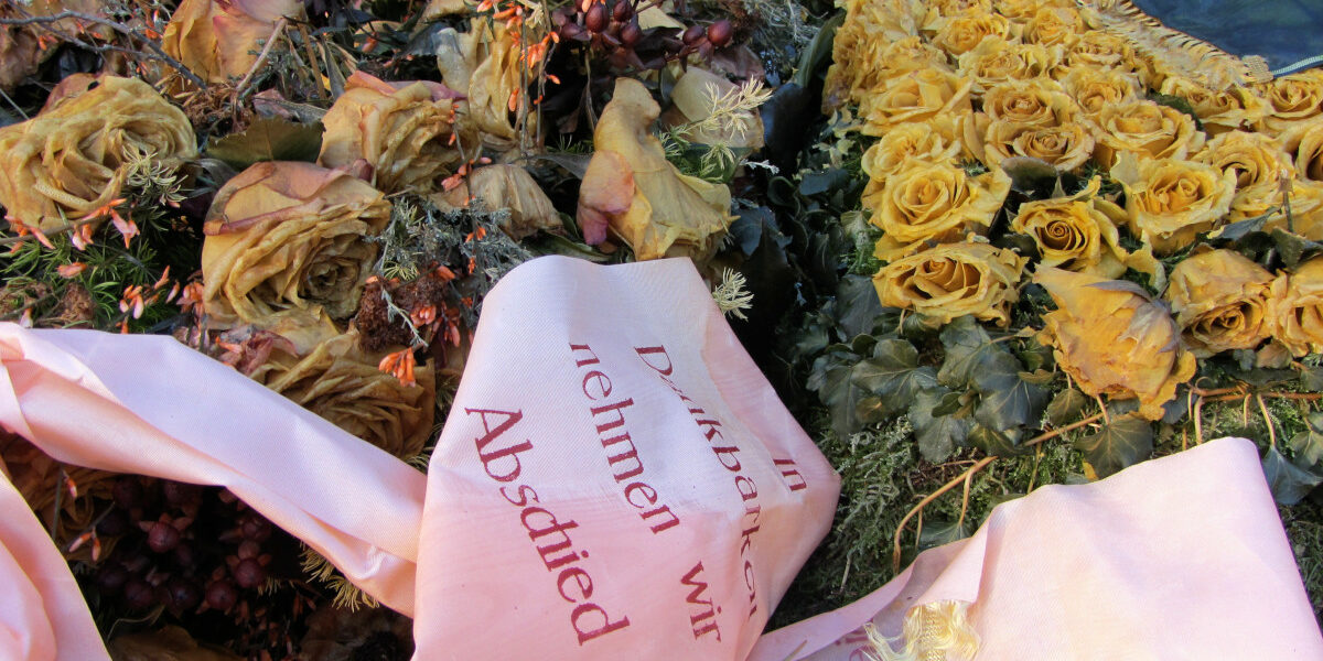Zu sehen ist ein verwelkter Blumenkranz mit einem Band, auf dem schwer lesbar "wir nehmen Abschied" steht.