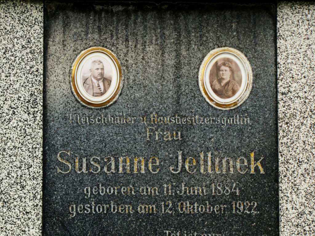 Ein Grabstein mit den schwarz-weiß Fotos eines Mannes und einer Frau. Dazu die Inschrift "Fleischhauer u. Hausbesitzersgattin Frau Susanne Jellinek"