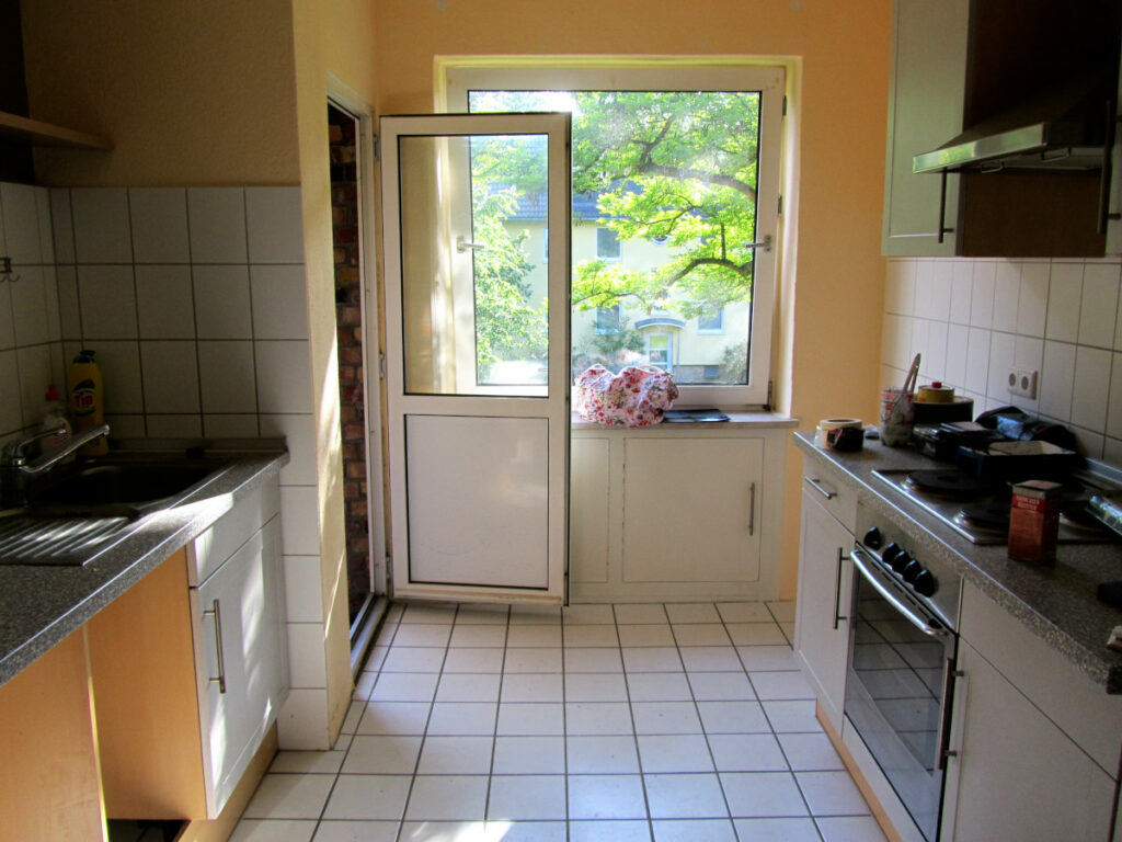Blick in eine Küche mit weißen Fliesen und Schränken und grauen Arbeitsplatten.