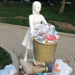 Ein überquellender Mülleimer, der aussieht wie ein Mädchen, das einen Korb hält. Drumherum zahlreicher Plastikmüll