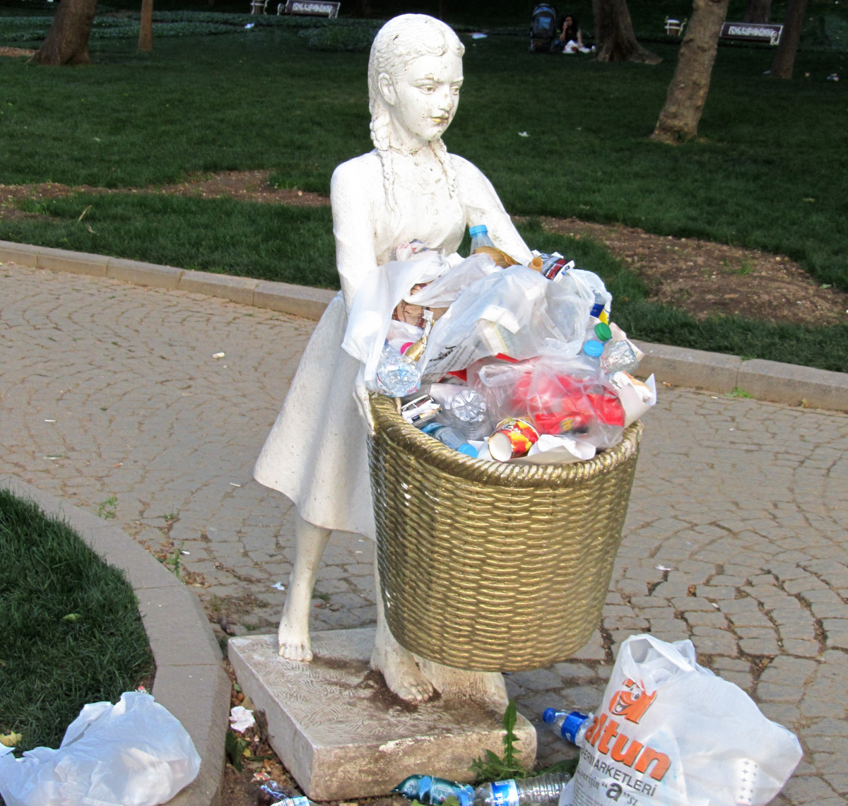 Ein überquellender Mülleimer, der aussieht wie ein Mädchen, das einen Korb hält. Drumherum zahlreicher Plastikmüll
